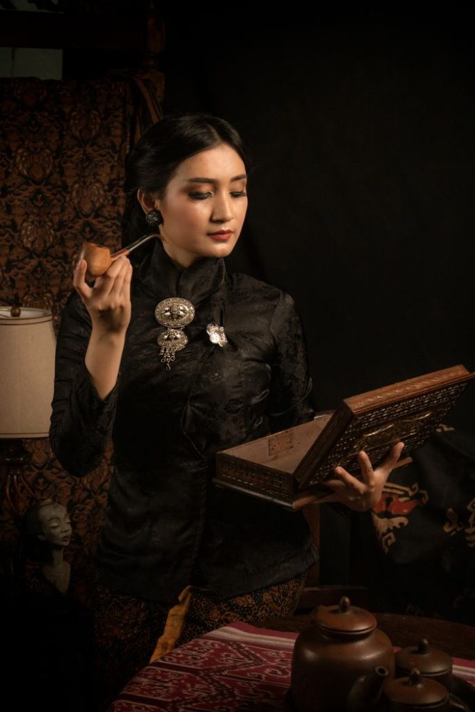 A Javanese woman in traditional attire holds a teapot and ornate box, reflecting Indonesian culture.